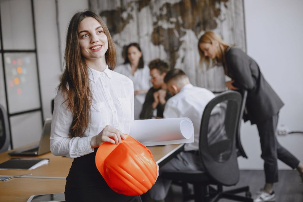 pexels-photo-4872030-4872030 Smiling businesswoman with a hard hat in a modern office setting, engaging with colleagues.